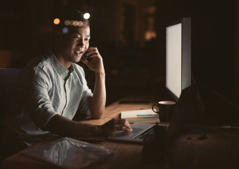 Smiling young Asian businessman talking on a cellphone while working in an office late at night with city lights glowing in the background
