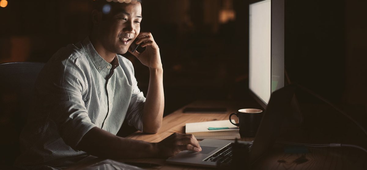 Smiling young Asian businessman talking on a cellphone while working in an office late at night with city lights glowing in the background